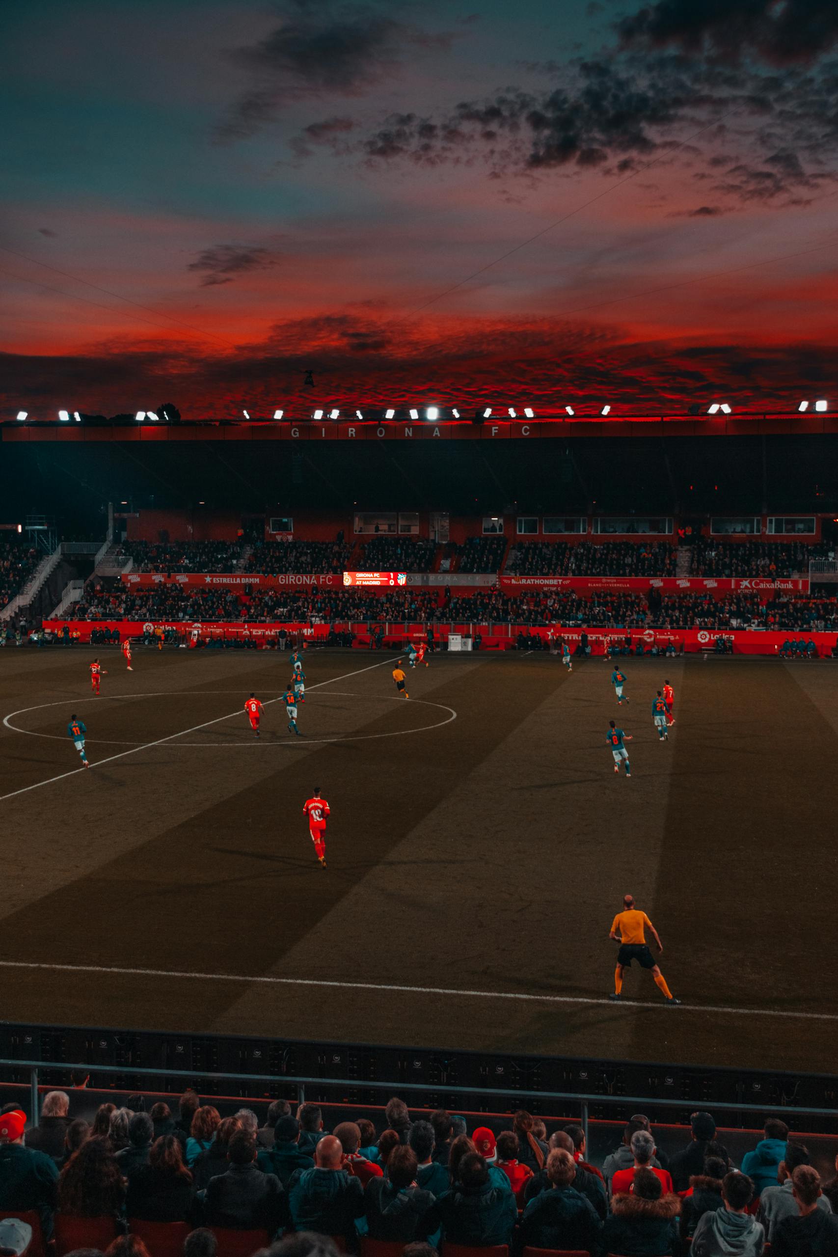 Wide-angle view of a soccer match taking place in a stadium at sunset with a vibrant sky.